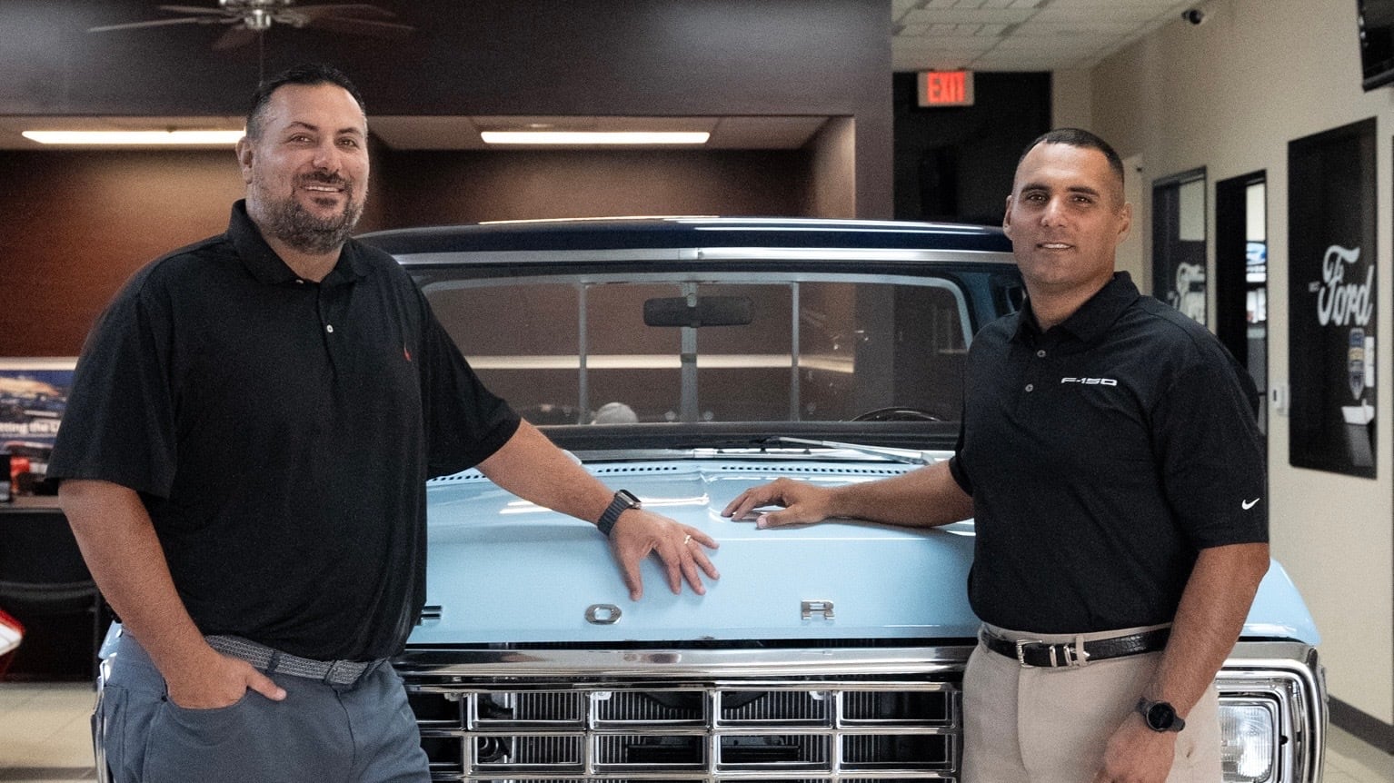 Two brothers dressed in black shirts pose in front of a vintage Ford truck inside an automotive dealership.